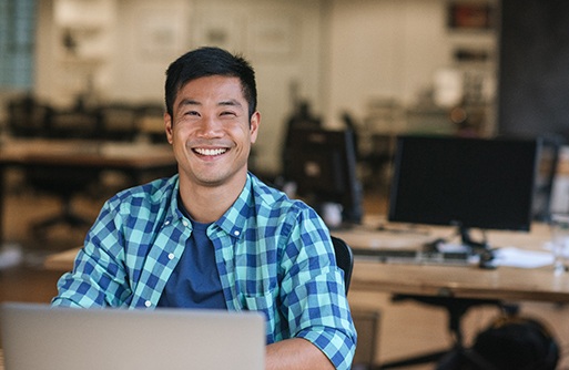 a smiling person sitting at a desk and working on a laptop